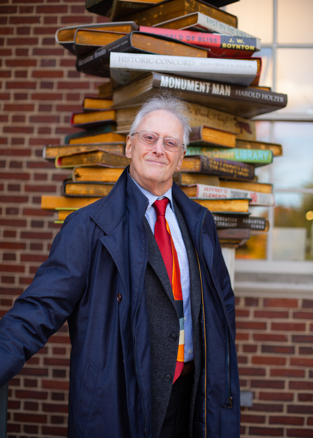 Dr. Gross standing in front of a sculpture depicting an irregular stack of large books with recognizable titles and authors on their spines.